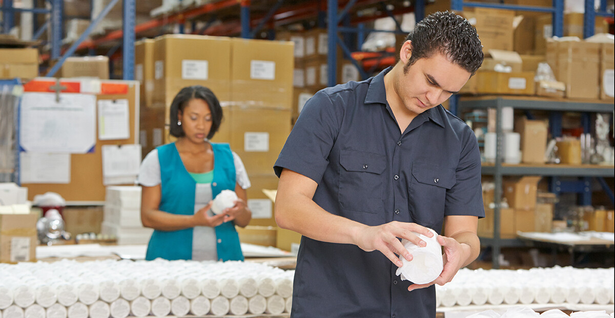 Zwei Beschäftigte prüfen verpackte Produkte an einem Arbeitstisch im Warenlager; im Hintergrund stehen hohe Regale und gestapelte Kartons.