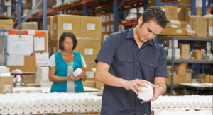 Zwei Beschäftigte prüfen verpackte Produkte an einem Arbeitstisch im Warenlager; im Hintergrund stehen hohe Regale und gestapelte Kartons.