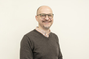 Porträt eines lächelnden Mannes mit Brille in kariertem Hemd und dunklem Pullover vor hellem Hintergrund.