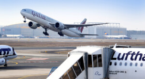Am Flughafen hebt ein Qatar-Airways-Jet ab, im Vordergrund steht ein Lufthansa-Flugzeug mit angedockter Fluggastbrücke.