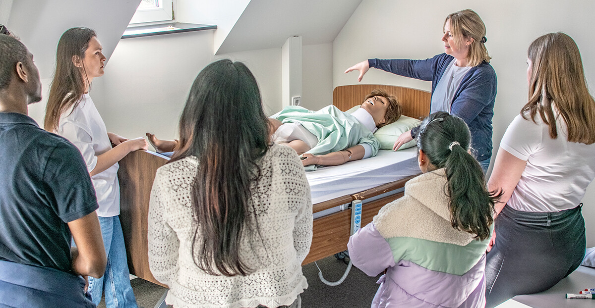 Eine Frau demonstriert an einer Pflegepuppe am Krankenhausbett eine Technik, mehrere Lernende stehen im Halbkreis und beobachten.