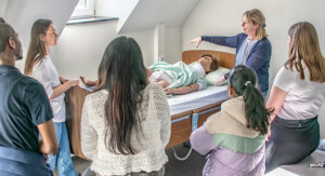 Eine Frau demonstriert an einer Pflegepuppe am Krankenhausbett eine Technik, mehrere Lernende stehen im Halbkreis und beobachten.