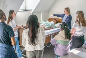 Eine Frau demonstriert an einer Pflegepuppe am Krankenhausbett eine Technik, mehrere Lernende stehen im Halbkreis und beobachten.