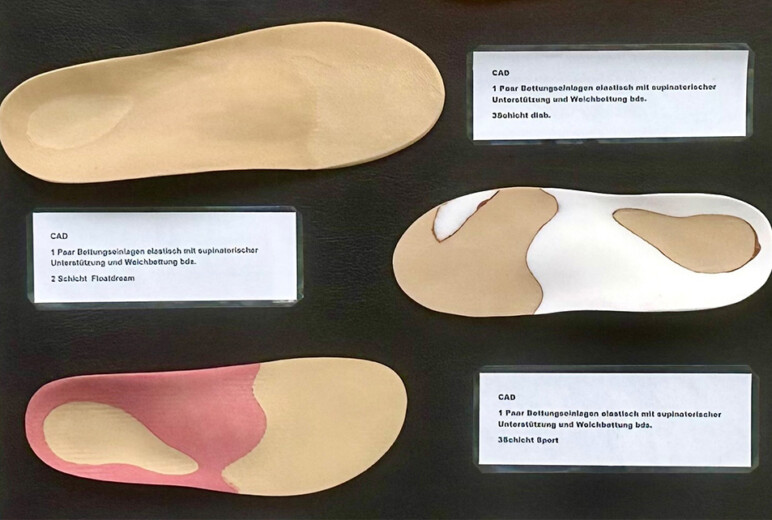 Nahaufnahme einer Präsentationswand mit mehreren orthopädischen Einlagen in unterschiedlichen Formen und Farben; kleine Beschriftungsschilder erklären die Modelle.