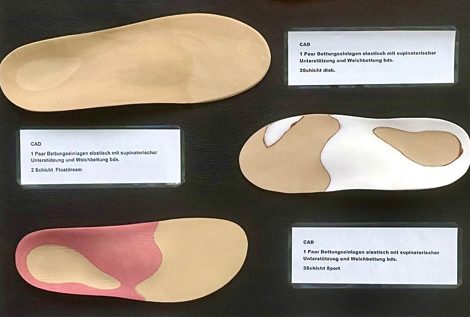 Nahaufnahme einer Präsentationswand mit mehreren orthopädischen Einlagen in unterschiedlichen Formen und Farben; kleine Beschriftungsschilder erklären die Modelle.