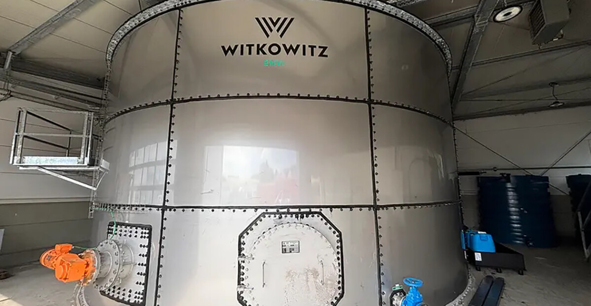 In einer Industriehalle steht ein großer, zylindrischer Stahltank mit dem Schriftzug „WITKOWITZ“; an den unteren Flanschen sind orange und blaue Armaturen montiert, darüber verlaufen Kabeltrassen und Lüftungselemente.