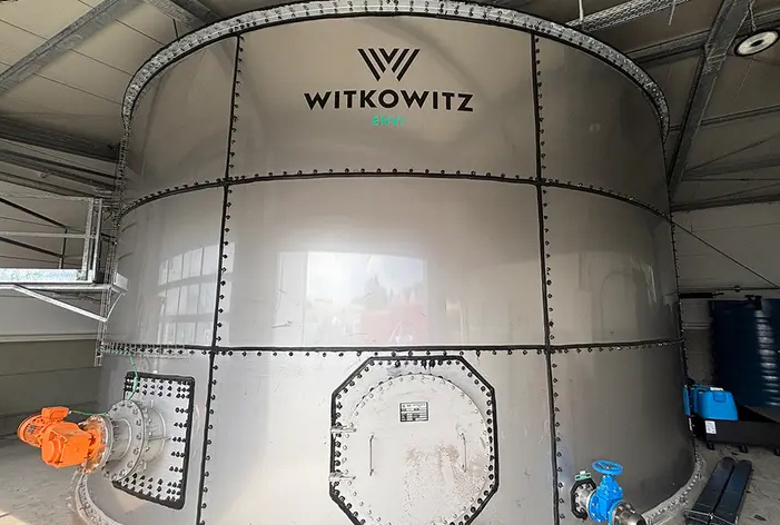 In einer Industriehalle steht ein großer, zylindrischer Stahltank mit dem Schriftzug „WITKOWITZ“; an den unteren Flanschen sind orange und blaue Armaturen montiert, darüber verlaufen Kabeltrassen und Lüftungselemente.