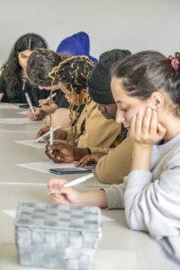 Teilnehmende sitzen in Reihe an Tischen und füllen konzentriert Arbeitsblätter aus; Stifte und Smartphones liegen davor.
