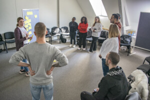Seminargruppe steht im Kreis. Eine Trainerin erklärt vor einer Pinnwand mit Post-its, die Teilnehmenden hören zu.