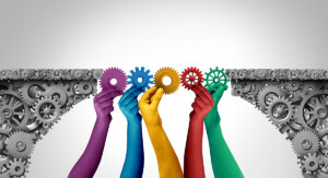 Mehrere farbige Hände halten Zahnräder, die zwei Zahnradflächen zu einer Brücke verbinden.
