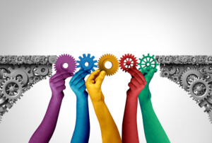 Mehrere farbige Hände halten Zahnräder, die zwei Zahnradflächen zu einer Brücke verbinden.