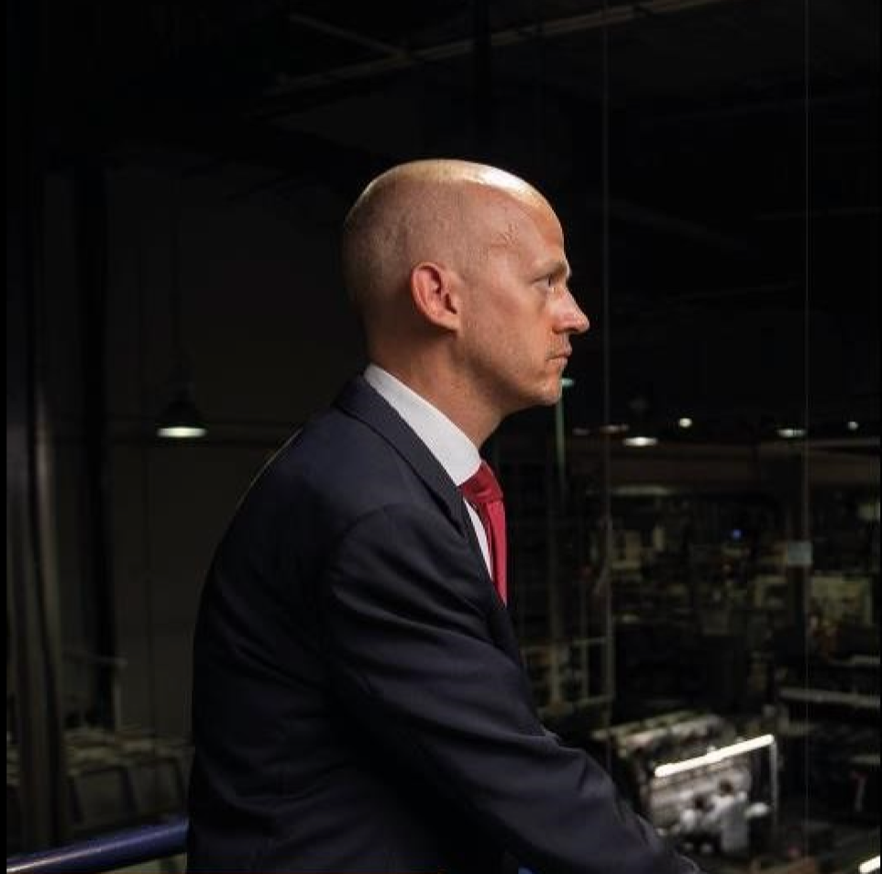 Ein Mann im Anzug blickt im Profil nach links in eine dunkle Industriehalle.