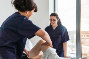 Eine Pflegekraft mobilisiert das gebeugte Bein einer Patientin am Bett, während eine Kollegin daneben steht und aufmerksam zusieht.