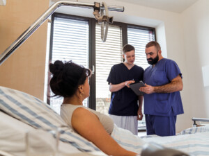 Zwei Pflegende mit Tablet stehen am Krankenbett und besprechen etwas mit einer Patientin, die halb aufgerichtet liegt.
