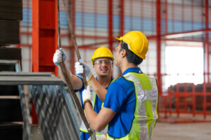 Zwei Bauarbeiter tragen Sicherheitshelme und Schutzwesten. Beide halten ein Seil und unterhalten sich. Die Szene findet auf einer Baustelle statt.