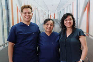 Drei Personen im Klinikflur, zwei in dunkelblauen Pflegeuniformen und eine in ziviler Kleidung, lächeln nebeneinander in die Kamera.