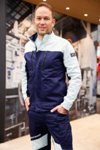 Junger Mann von vorne mit den Händen in den Taschen in Hose und Jacke von Kübler an einem Stand auf der A+A.