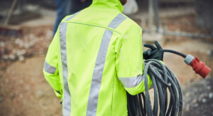 Person von hinten mit Warnweste und schwarzen Handschuhen von hinten, die einen gerollten Wasserschlauch mit dem rechten Unterarm und der rechten Hand hält. Man sieht weder Kopf noch Beine.